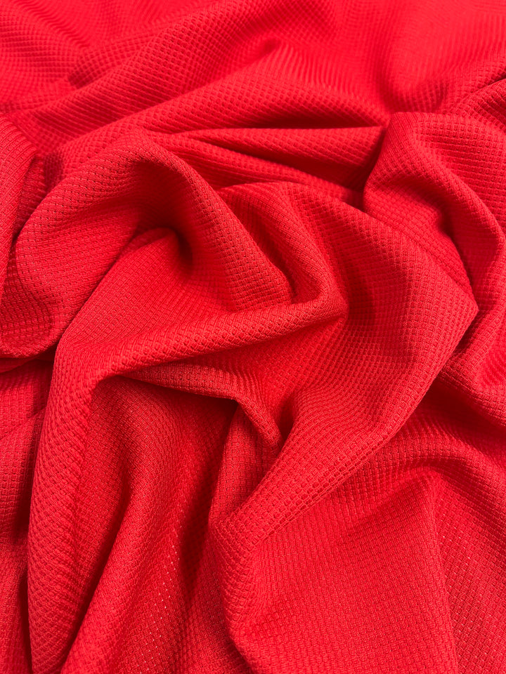 Close-up image of bright red textured fabric, Super Cheap Fabrics' Waffle Knit - Red Alert - 170cm, with a slightly wrinkled appearance. The material appears soft and displays a small, repeating pattern reminiscent of honeycomb fabric. The folds create shadow and depth, adding to the three-dimensional visual interest of the cloth.