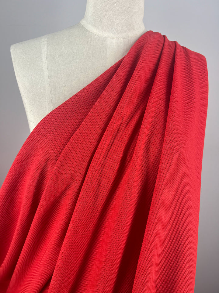 A mannequin draped with vibrant red **Super Cheap Fabrics Waffle Knit - Red Alert - 170cm** covers the left shoulder. The honeycomb fabric features a subtle, small-scale pattern that adds a three-dimensional appearance. The background is a plain light gray, highlighting the color and texture of the fabric.