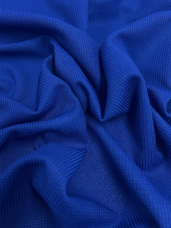A close-up view of a deep blue, textured fabric with a subtle grid pattern. The Waffle Knit - Cobalt - 170cm by Super Cheap Fabrics is softly draped and gathered, creating gentle folds and shadows that enhance its three-dimensional effect.