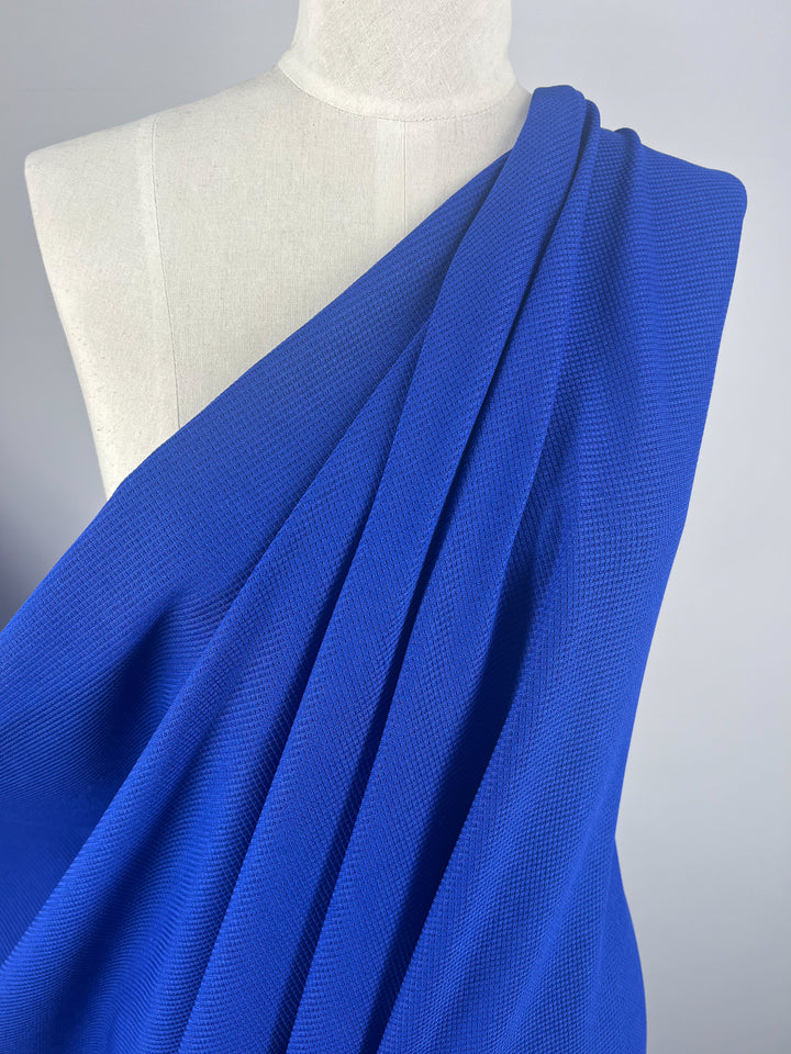 A white mannequin is draped with Super Cheap Fabrics' Waffle Knit - Cobalt - 170cm, which cascades over one shoulder. The textured, slightly ribbed appearance of the material adds a three-dimensional effect. The plain and neutral background accentuates the vibrant color of the fabric.