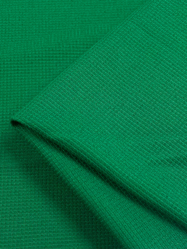 Close-up image of a folded piece of Super Cheap Fabrics Waffle Knit - Abundant Green - 170cm, showing its weave and detailed pattern. The fabric appears soft and slightly stretchy, with a visible grid-like texture. The fold creates a clean edge, emphasizing the material's three-dimensional appearance and quality.