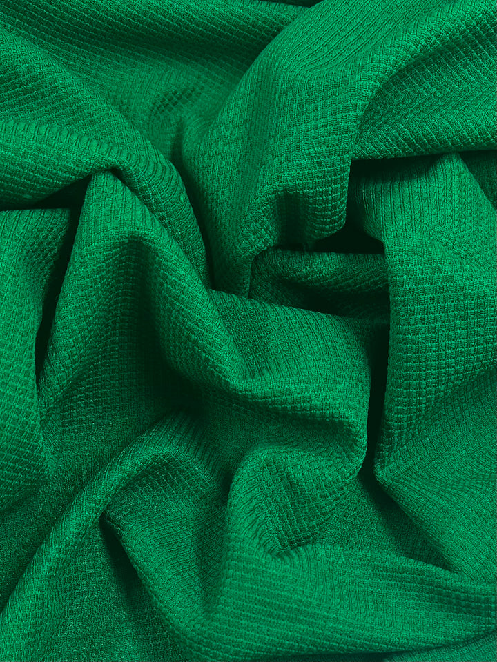 Close-up of a green textured Waffle Knit - Abundant Green - 170cm fabric from Super Cheap Fabrics with a three-dimensional appearance. The fabric is draped and folded, creating shadows and highlights that enhance its textured look. The deep green color is consistent throughout the cloth.
