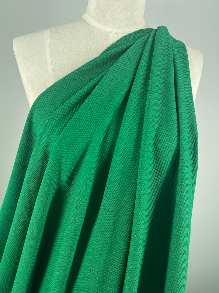 A draped piece of bright green Waffle Knit - Abundant Green - 170cm from Super Cheap Fabrics is elegantly spread over the shoulder of a white mannequin. The textured pattern creates a three-dimensional appearance with flowing folds, adding to its graceful look. The background remains plain and unobtrusive.