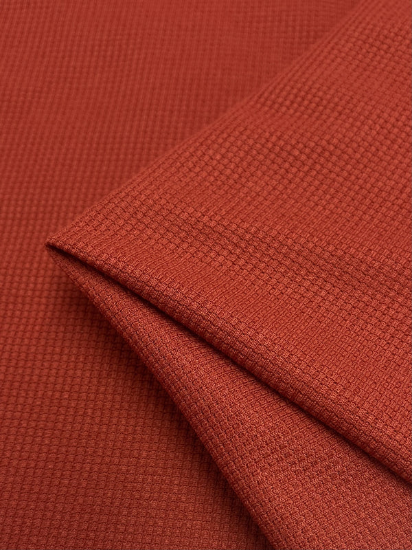 Close-up image of folded rust-colored fabric with a subtle, textured pattern. The material appears to be lightweight and finely woven, featuring a unique three-dimensional appearance reminiscent of Super Cheap Fabrics' Waffle Knit - Rooibos Tea - 170cm.