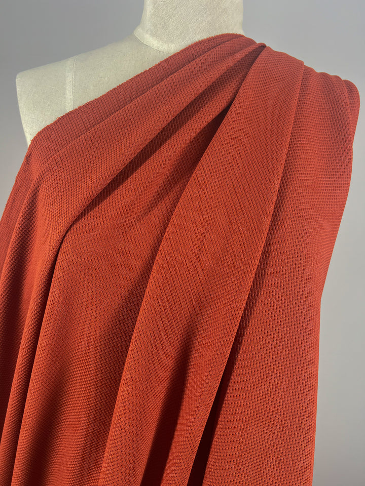 A coral-red Waffle Knit - Rooibos Tea - 170cm fabric from Super Cheap Fabrics with a textured, honeycomb-like pattern is draped over a mannequin. The fabric covers the left shoulder and cascades smoothly, highlighting its soft and flowy nature. The background is plain and gray, emphasizing the fabric’s three-dimensional appearance.