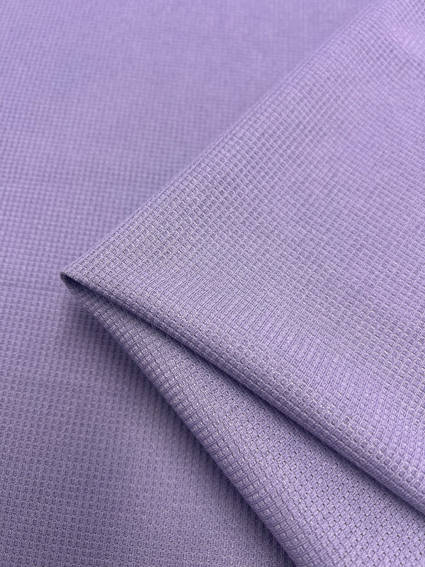 A close-up image of two overlapping pieces of light purple waffle fabric with a subtle grid-like texture. The fabric appears to be neatly folded, showcasing its smooth surface and fine pattern. This is the Waffle Knit - Lilac - 170cm by Super Cheap Fabrics.