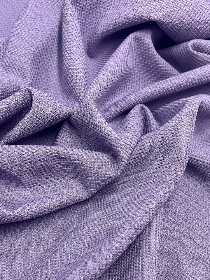 A close-up of smooth, crumpled Waffle Knit - Lilac - 170cm fabric by Super Cheap Fabrics in a light lavender color. The material has a subtle, textured grid pattern, creating a soft and elegant appearance. The folds and creases enhance the fabric's tactile quality.