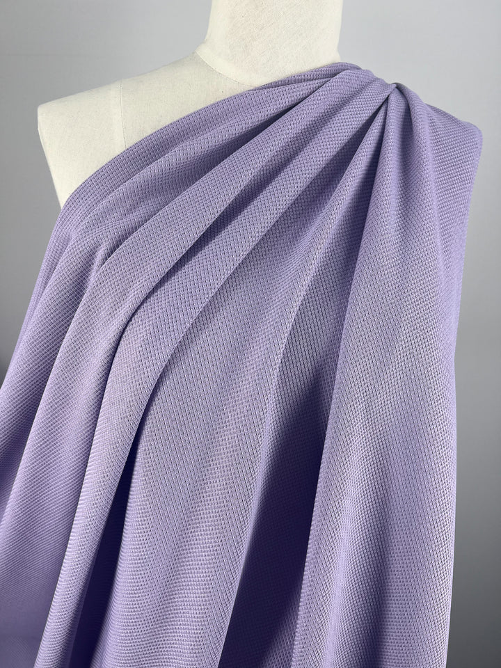 Close-up of a mannequin draped in a soft, pastel purple fabric. The delicate Waffle Knit - Lilac - 170cm from Super Cheap Fabrics features an intricate pattern and is elegantly wrapped around the mannequin's shoulders, creating gentle folds and pleats. The background is a neutral gray.