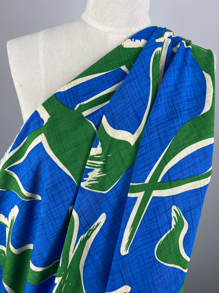 A white dress form is draped with lightweight fabric featuring a blue background. The vibrant Bamboo Rayon - Dance Indigo - 150cm by Super Cheap Fabrics is adorned with abstract green and white designs, creating a dynamic and colorful pattern. Perfect for beginner-friendly sewing, the dress form stands against a plain gray backdrop.