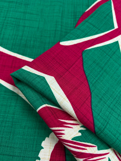 Close-up image of a folded Super Cheap Fabrics Bamboo Rayon - Dance Pepper Green - 150cm with a vibrant pattern, featuring interlocking crimson red and white shapes on a deep green background. The texture of this lightweight fabric appears lightly crosshatched, adding dimension to the vivid colors and geometric design, perfect for home decor.