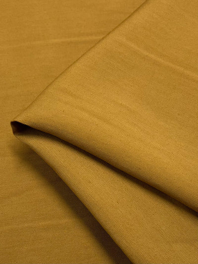 A close-up view of a mustard-colored suiting fabric folded neatly, showcasing its smooth texture and fine weave. The lightweight **Suiting - Honey Mustard - 140cm** by **Super Cheap Fabrics** appears to be slightly matte, with no visible patterns or designs.