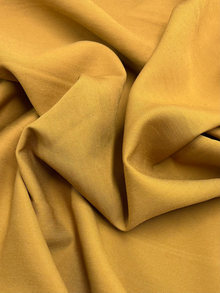 A close-up image of Super Cheap Fabrics' Suiting - Honey Mustard - 140cm. The cloth is slightly crumpled, showcasing its texture and sheen under soft lighting. The folds create gentle shadows, highlighting the material's smooth and continuous surface, characteristic of fine light weight suiting.