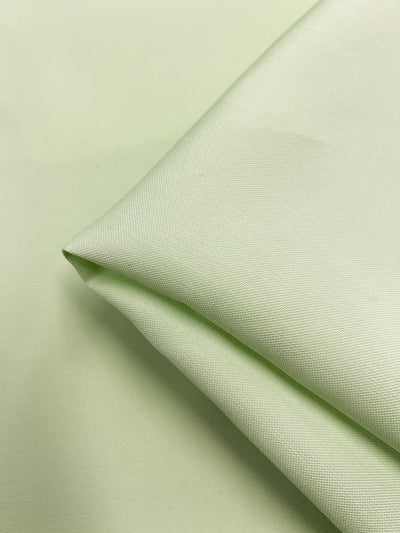 A close-up view of a light green piece of fabric, neatly folded at one corner. The Suiting - Ambrosia - 140cm by Super Cheap Fabrics appears smooth and has a fine texture. The background is also light green, similar to the fabric color, creating a uniform and serene appearance.