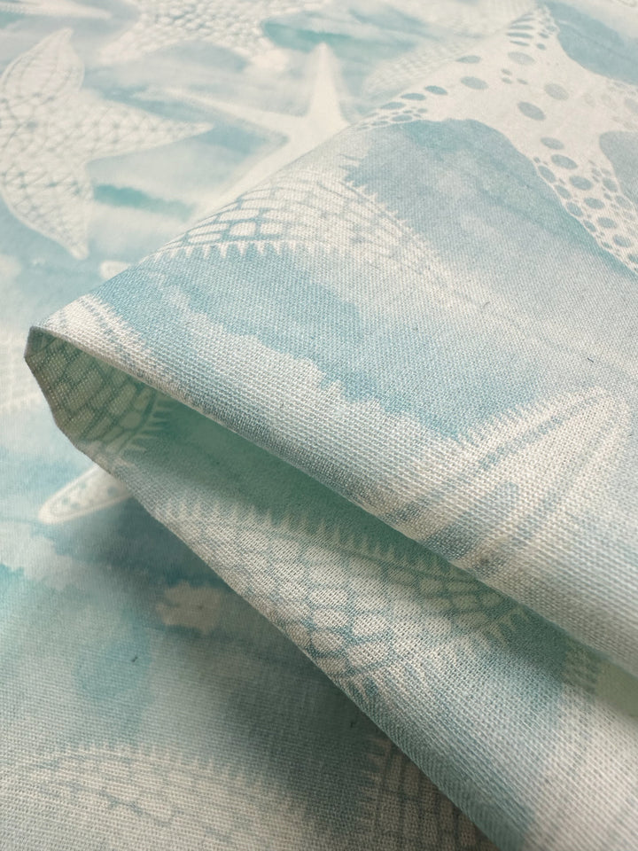 A close-up of the "Under The Sea" light blue printed cotton fabric from Super Cheap Fabrics, featuring starfish and distressed textures. The 150cm width natural cotton fibers are folded at an angle, elegantly highlighting the pattern and tactile allure.