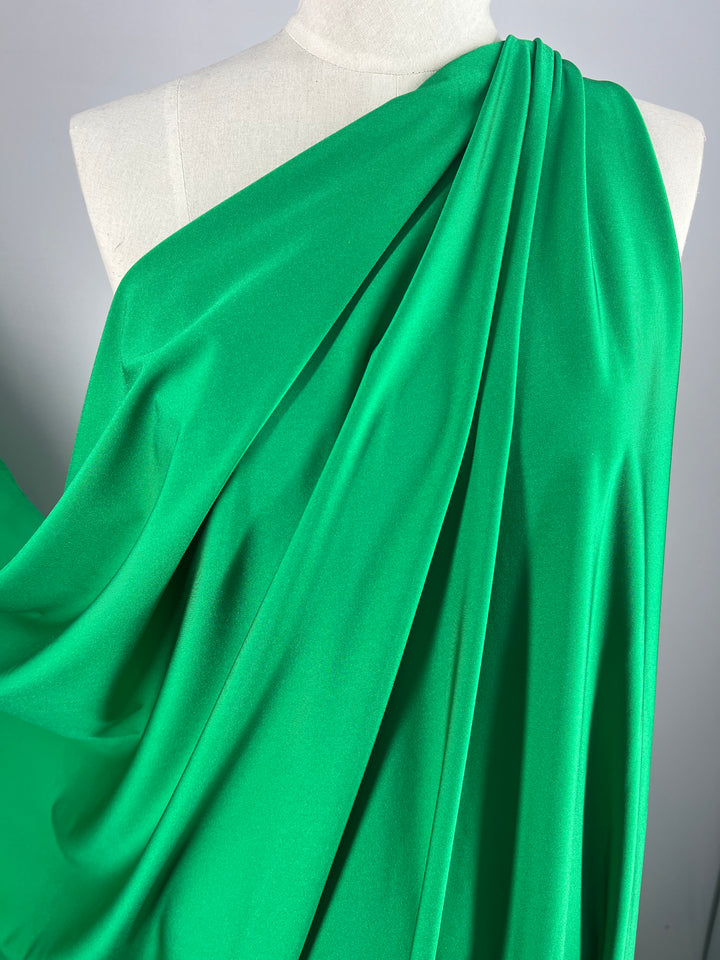 A close-up of a mannequin draped in vibrant green Nylon Lycra - First Tee - 150cm by Super Cheap Fabrics. The fabric is arranged to cover one shoulder, creating flowing and elegant folds. The texture appears smooth and silky with a luxurious four-way stretch, highlighting its rich color ideal for swimwear and dancewear.