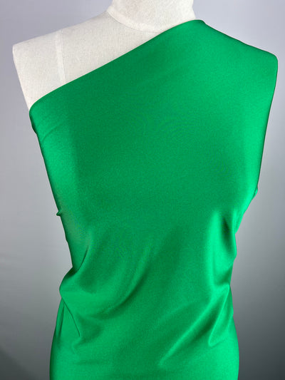A close-up of a mannequin dressed in a bright green, one-shoulder garment made from Super Cheap Fabrics' Nylon Lycra - First Tee - 150cm fabric. The smooth material has four-way stretch and drapes elegantly over the form, highlighting the vibrant color and sleek design. The plain, light-colored background ensures the garment stands out.