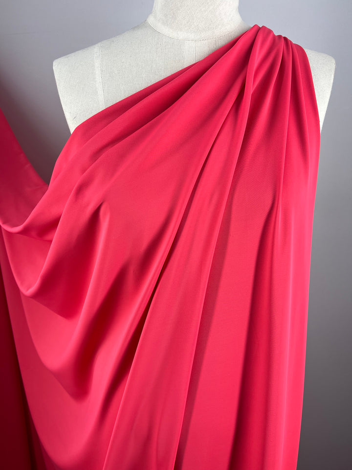 A close-up photograph shows a dress form draped with flowy, bright pink Nylon Lycra - Azalea - 160cm fabric from Super Cheap Fabrics. The material, known for its four-way stretch, is gathered over one shoulder in a way that creates soft folds and a sense of movement. The background is a plain, light gray color, emphasizing the vibrant hue of the fabric.