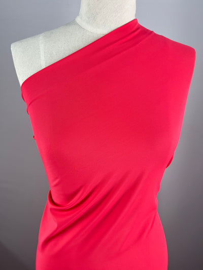 A red dress on a mannequin with an asymmetrical one-shoulder design in Super Cheap Fabrics' Nylon Lycra - Azalea - 160cm. The material drapes smoothly, showcasing a sleek and elegant style. The plain gray background highlights the bright red color of the dress, making it perfect for swimwear or evening occasions with its four-way stretch.
