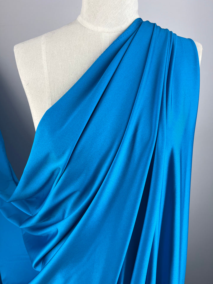 A close-up of a draped mannequin adorned with vibrant Super Cheap Fabrics Nylon Lycra - Vivid Blue - 155cm. The swimwear fabric, which has a smooth and shiny texture, cascades gracefully over the mannequin, forming gentle, elegant folds around the shoulder area. The background is plain and unobtrusive.