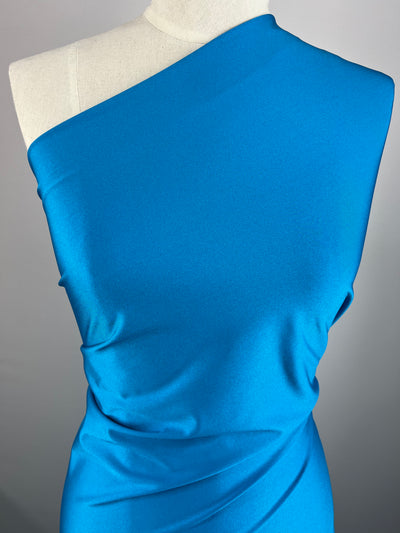A torso mannequin displays a vibrant blue, one-shoulder dress made from Super Cheap Fabrics' Nylon Lycra - Vivid Blue - 155cm fabric. The dress has a smooth texture with gentle draping effects on one side. The background is plain, allowing the vivid blue color of the stretchable fabric to stand out.