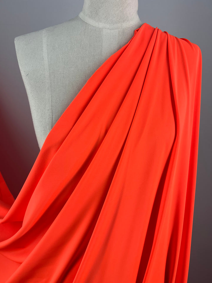 A mannequin draped with vibrant Super Cheap Fabrics' Nylon Lycra - Fluro Orange - 160cm fabric, showcasing its flow and texture. The swimwear fabric is arranged across the shoulder and chest, displaying folds and pleats that highlight its smooth and flexible nature. The background is plain, focusing attention on the fabric.