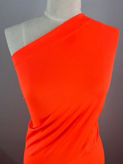 A close-up view of a mannequin dressed in a bright red, one-shoulder garment made from **Super Cheap Fabrics' Nylon Lycra - Fluro Orange - 160cm**. The smooth and form-fitting swimwear fabric showcases a minimalistic and elegant design. The background is a plain gray, putting the focus entirely on the striking red garment.