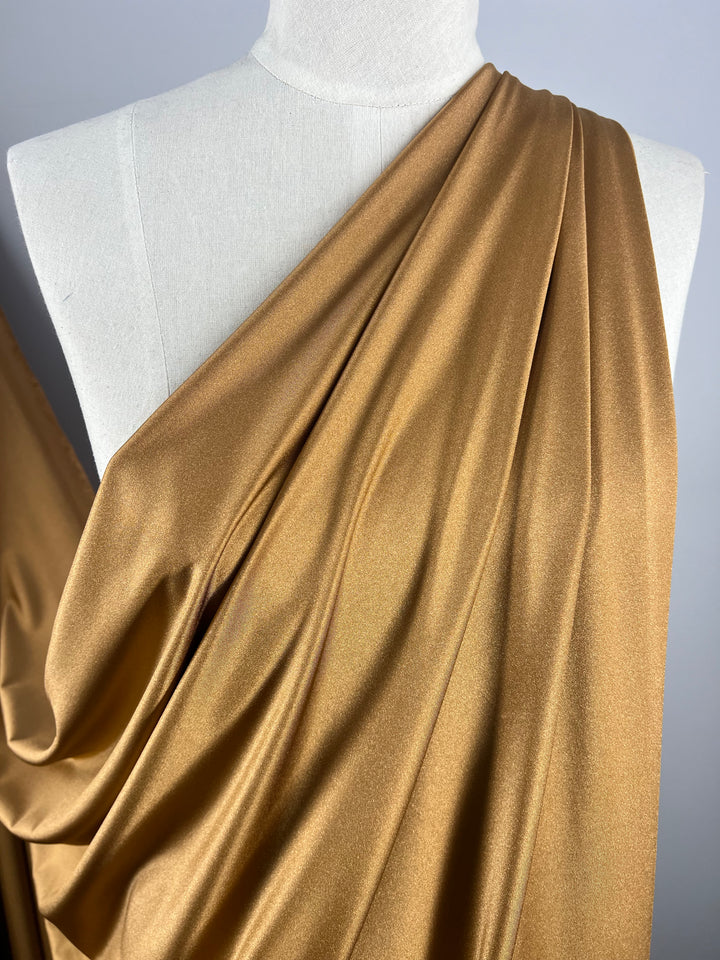 A piece of golden fabric, reminiscent of Super Cheap Fabrics' Nylon Lycra - Tawny Birch - 155cm, is draped elegantly on a white dress form. The smooth, shimmery texture is arranged in loose folds, showcasing its rich color and high-quality sheen. The background remains a plain, light gray.
