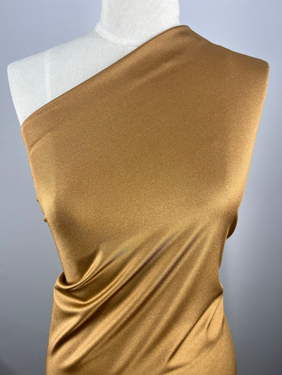 A close-up of a mannequin draped in Super Cheap Fabrics' Nylon Lycra - Tawny Birch - 155cm. The material has a sheen and is styled in an asymmetrical, one-shoulder design. The background is a plain, light grey color, which makes the golden fabric stand out.