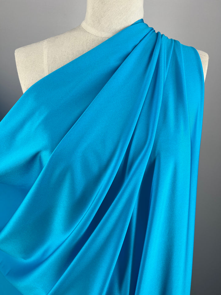A close-up of a mannequin draped in vibrant blue Super Cheap Fabrics Nylon Lycra - Horizon Blue - 155cm fabric. The material is gathered and styled elegantly over one shoulder, showcasing smooth, flowing folds and a soft texture. The neutral backdrop allows the bright blue, synonymous with swimwear fabric, to stand out prominently.
