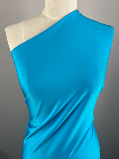 A mannequin is dressed in a one-shoulder, bright blue dress crafted from Super Cheap Fabrics Nylon Lycra - Horizon Blue - 155cm. The material drapes smoothly across the torso, creating a sleek and elegant appearance. The background is plain and neutral, ensuring the dress stands out prominently.