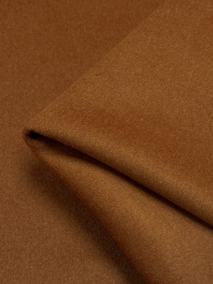 Close-up of a folded piece of **Super Cheap Fabrics Wool Cashmere - Leather Brown - 150cm**. The texture appears soft and smooth, similar to thick wool cashmere fabric, and the edges are neatly folded to form a sharp corner. The lighting highlights the fabric's texture and color variations subtly.