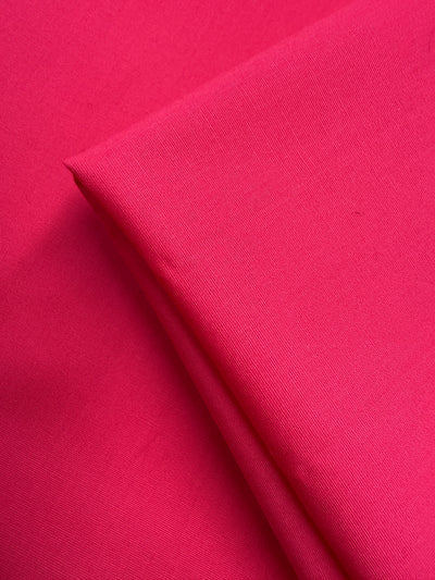 A close-up of Super Cheap Fabrics' Plain Cotton - Fuchsia - 150cm shows two pieces, one folded atop the other, highlighting their smooth, slightly textured surface.