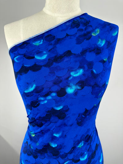 A mannequin is draped in a vivid blue, one-shoulder garment made from medium-weight Printed Lycra - Scales - 150cm by Super Cheap Fabrics and adorned with a shimmering pattern resembling fish scales. The material creates an iridescent effect, reflecting different shades of blue and hints of green. The background is plain and light-colored.