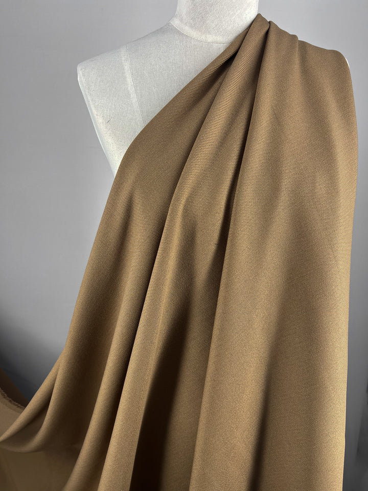 Close-up of a mannequin draped with a large piece of light brown, Twill Suiting - Aztec - 138cm from Super Cheap Fabrics. The fabric covers the mannequin’s shoulder and extends downward, exhibiting a smooth texture and soft folds. The background is neutral, accentuating the color and texture of the fabric.
