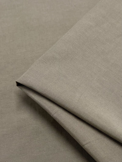 A close-up photo of a neatly folded piece of medium-weight, light gray Italian Linen Blend - Tannin - 150cm by Super Cheap Fabrics. The texture appears smooth and slightly matte, with subtle linear patterns. The fabric is layered over a larger piece of the same material, showing its folds and edges beautifully.