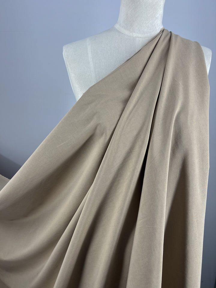 A beige Italian linen fabric, Linen Blend - Tannin - 150cm by Super Cheap Fabrics, is draped elegantly over a white mannequin torso against a solid gray background. The medium weight linen fabric has soft folds and a smooth texture, giving it a sense of movement and fluidity.