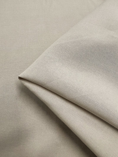 Close-up view of a folded piece of light beige, lightweight fabric with a smooth texture. The material, likely Linen Blend - Cement - 150cm from Super Cheap Fabrics, is slightly reflective, and the fold creates a sharp diagonal line across the image.