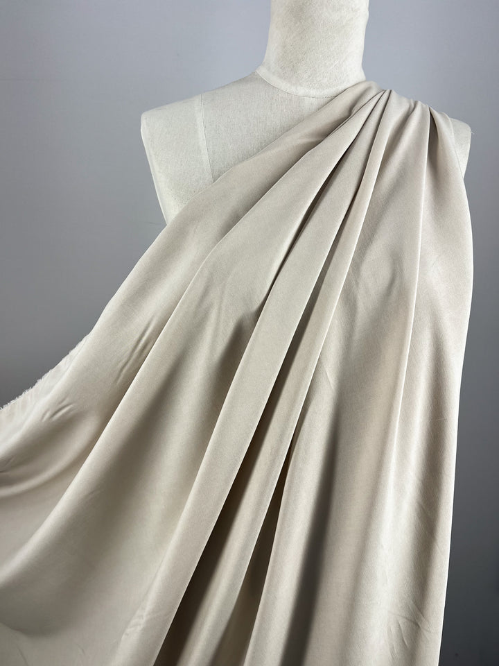 A mannequin is draped with a large piece of Linen Blend - Cement - 150cm by Super Cheap Fabrics, creating soft, flowing folds. The lightweight fabric has a smooth texture and hangs elegantly over the mannequin's shoulders and chest against a plain gray background.