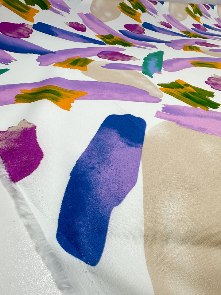 The Printed Cotton - Petals - 150cm from Super Cheap Fabrics is a vivid textile featuring abstract watercolor brushstrokes in blue, purple, green, orange, and beige against a white background. Made from natural cotton fibers, this versatile fabric displays diverse shapes and sizes for an energetic and colorful design.