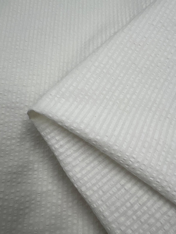 A close-up photo of neatly folded Seersucker Cotton - Off White - 155cm fabric with a subtle textured pattern of small squares and ridges. The medium weight cotton fabric from Super Cheap Fabrics appears soft and clean, with the pattern creating a visually interesting detail.