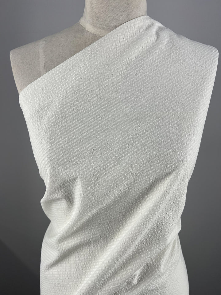 A mannequin draped with Super Cheap Fabrics' Seersucker Cotton - Off White - 155cm. The off white fabric is wrapped asymmetrically around the mannequin, covering one shoulder and leaving the other exposed. The backdrop is a plain, gray background.