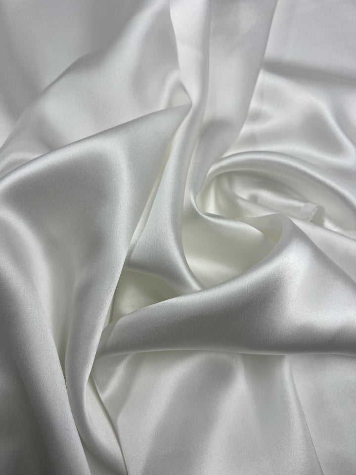 Close-up of a smooth, white Silk Satin - Off White - 138cm fabric by Super Cheap Fabrics, displaying its lustrous and glossy texture. The luxurious material is gathered and gently folded, creating soft curves and flowing lines that highlight its silky appearance, perfect for crafting elegant dressy tops.