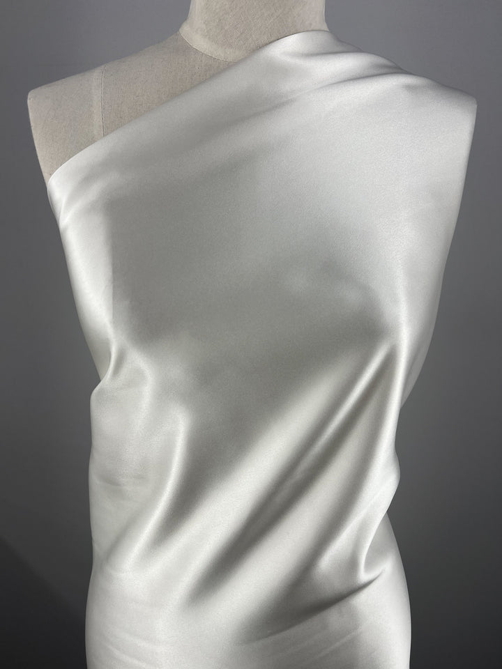 A close-up of a mannequin draped in Super Cheap Fabrics' Silk Satin - Off White - 138cm. The material is arranged in an elegant, asymmetrical style, highlighting its glossy, reflective surface and soft, flowing texture. The background is plain and dark, emphasizing the sheen of the satin.