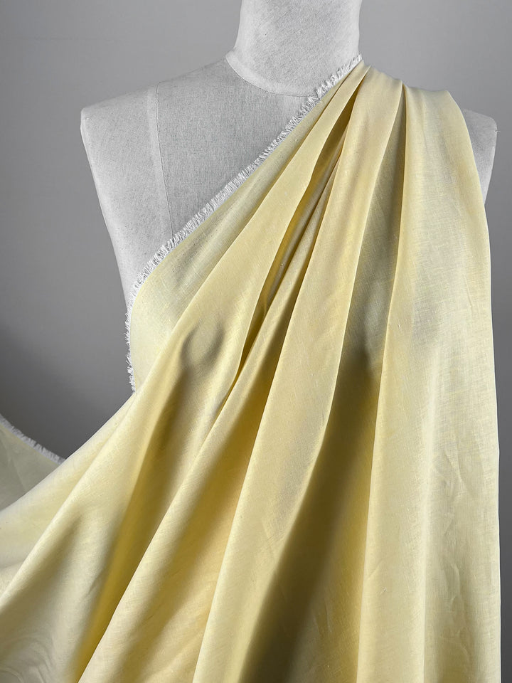 A Linen Voile - French Vanilla - 155cm by Super Cheap Fabrics is elegantly draped over a white mannequin, demonstrating its soft texture and fluidity. The material has a light sheen and is fringed along one edge, adding a delicate detail to the overall appearance. The background is a neutral gray.