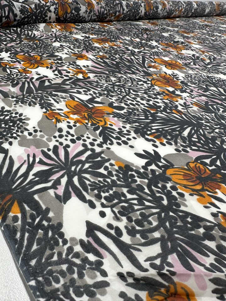 A detailed view of the Printed Silk Voile - Garzen from Super Cheap Fabrics reveals an abstract floral pattern in black, gray, orange, and pink. This design incorporates different flower and leaf shapes, producing a layered appearance on its extra lightweight material that is elegantly displayed on a flat surface.