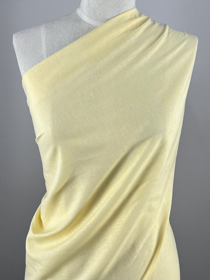A light yellow, lightweight fabric from Super Cheap Fabrics, Linen Voile - French Vanilla - 155cm, is draped over a torso mannequin, covering the mannequin from the left shoulder down to the lower right side. The cotton material has a subtle texture and a gentle, flowing appearance. The background is plain and gray.