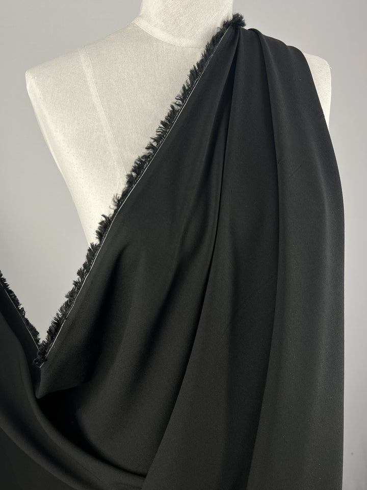 A close-up image of a mannequin draped with Super Cheap Fabrics' Premium Viscose Suiting - Raven - 130cm. The medium to heavy weight fabric has a slightly textured edge and falls smoothly over the mannequin, showcasing its flow and drape. The background is a plain light gray, which contrasts with the dark fabric.