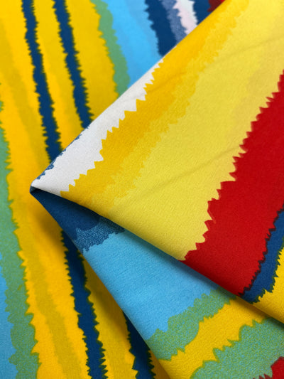 Close-up of colorful Deluxe Print - Beachside - 155cm fabric with wavy, stripe patterns by Super Cheap Fabrics. The fabric features bold, jagged lines in vibrant hues including yellow, red, blue, and green. Part of the designer fabric is folded, showcasing overlapping designs and creating a dynamic, eye-catching visual.