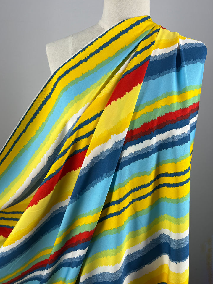A close-up of a mannequin draped with Super Cheap Fabrics' Deluxe Print - Beachside - 155cm featuring vibrant, wavy horizontal stripes in yellow, blue, red, white, and teal. The colorful design creates a lively, dynamic effect ideal for any designer fabric collection.