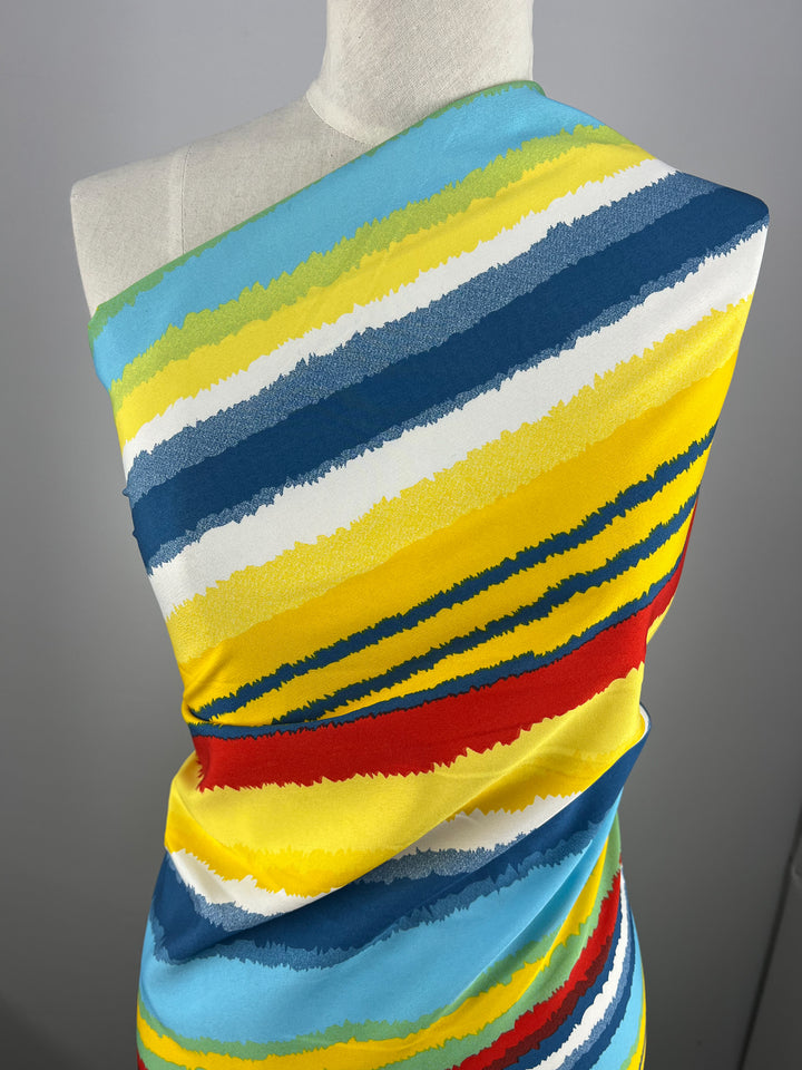 A white mannequin is draped with a colorful, versatile polyester fabric featuring bold horizontal stripes in blue, yellow, red, white, and light blue. The Deluxe Print - Beachside - 155cm by Super Cheap Fabrics wraps around the torso, showcasing the vibrant design. The background is plain and gray.